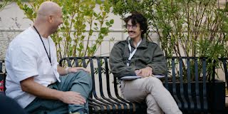 Two individuals are in discussion in the garden, seated at a desk.