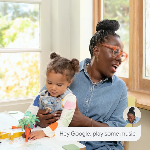 A parent holds their child and says 'Hey Google, play some music' without interrupting playtime. 
