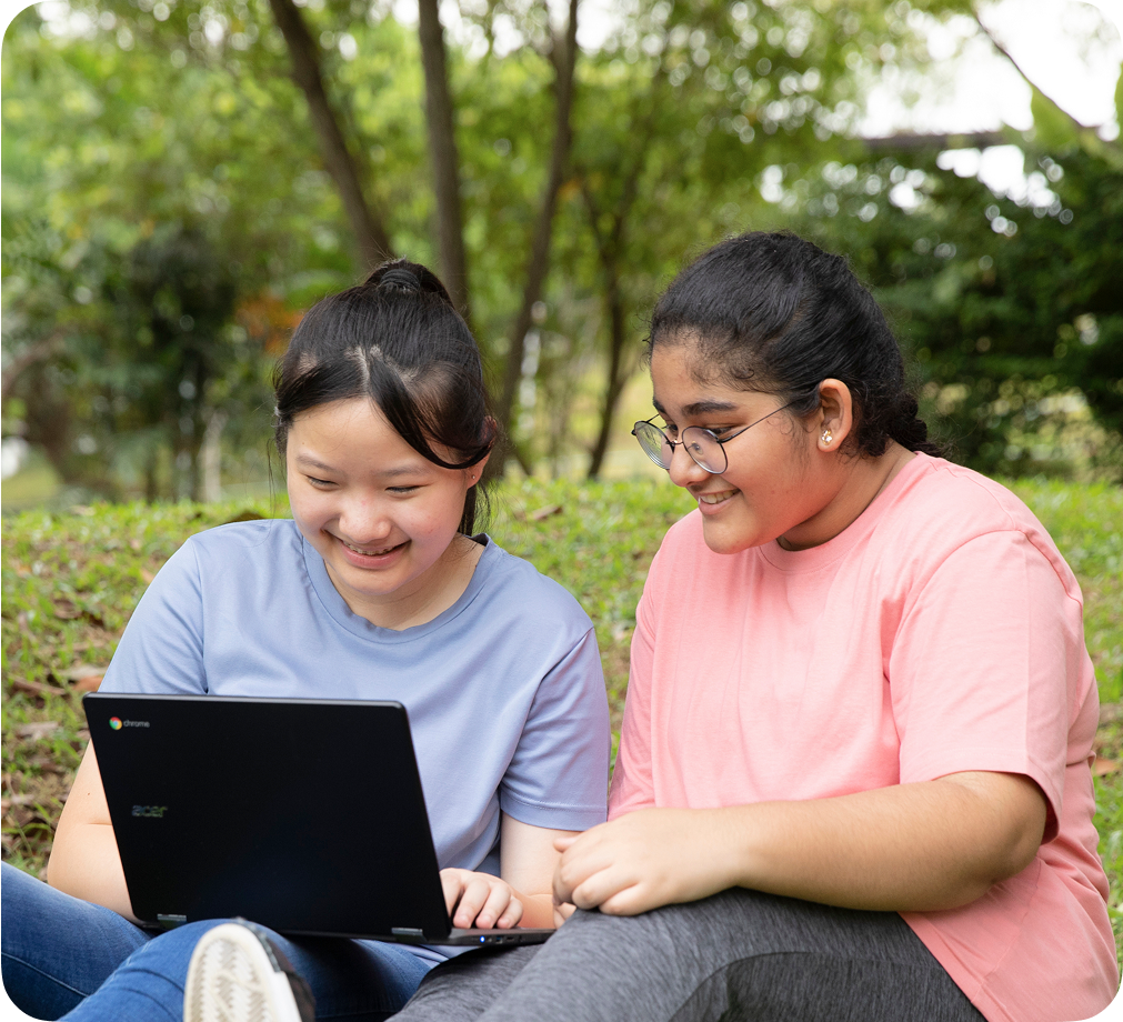 Dua anak yang tersenyum duduk bersebelahan di luar dan bekerja bersama menggunakan perangkat Chromebook.